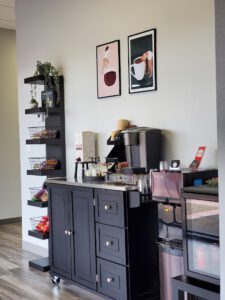 A coffee station with a black cabinet, coffee machine, cups, condiments, snacks on shelves, mini fridge, and two art prints on the wall above. - Modish Salon Professional