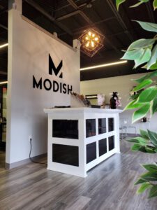 A modern salon reception area with a white desk, "MODISH" sign on the wall, geometric light fixture, and greenery in the foreground. - Modish Salon Professional