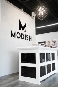 Modern reception desk with "MODISH" logo on the white wall behind, under geometric pendant light in a contemporary office or salon setting. - Modish Salon Professional