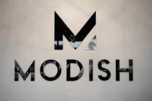 A black, geometric letter "M" and the word "MODISH" are mounted on a light gray wall. - Modish Salon Professional