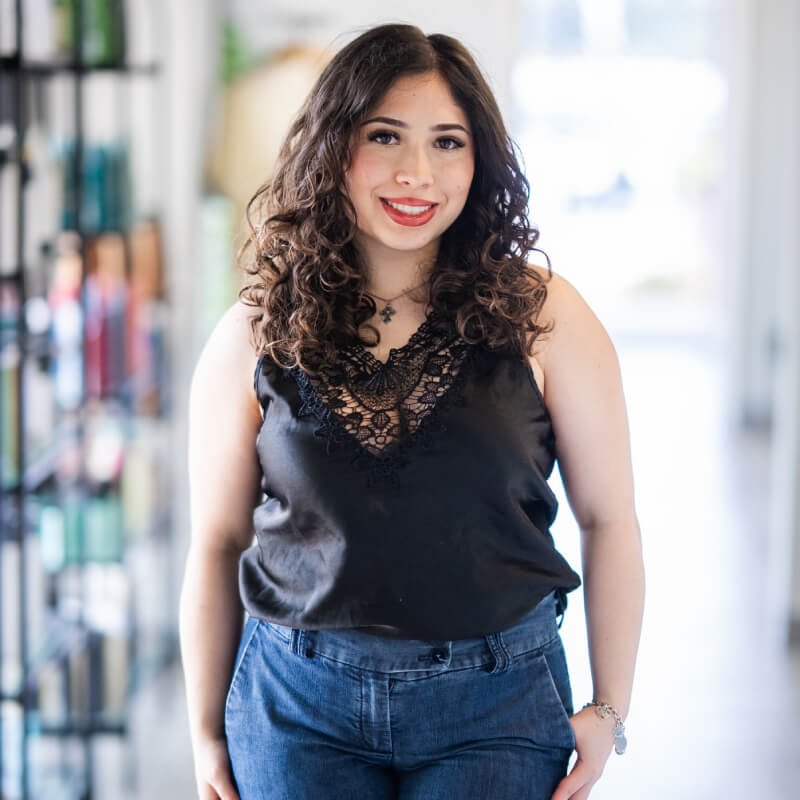 A smiling woman with long hair stands in front of shelves with various hair products, wearing a floral dress. -Pure Salon, Ankeny, Johnston, & Indianola, IA