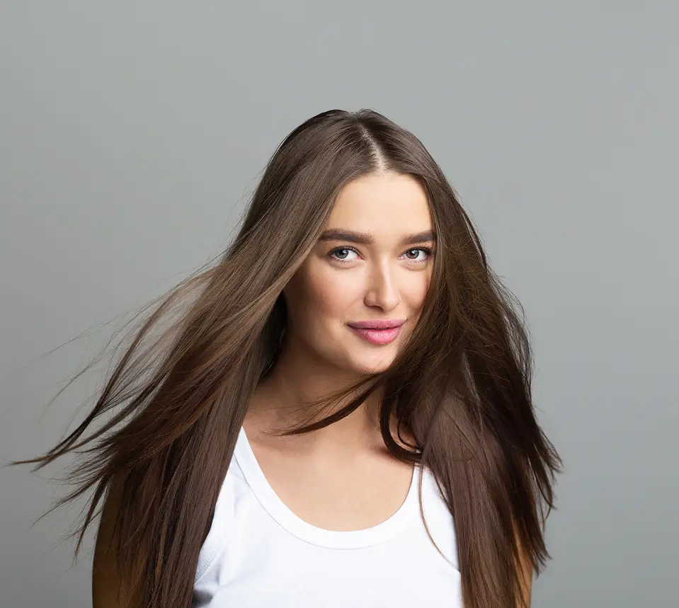 Woman with long straight brown hair, freshly styled in a salon, wearing a white tank top. She stands against a plain gray background, looking at the camera and smiling slightly. - Modish Salon Professional