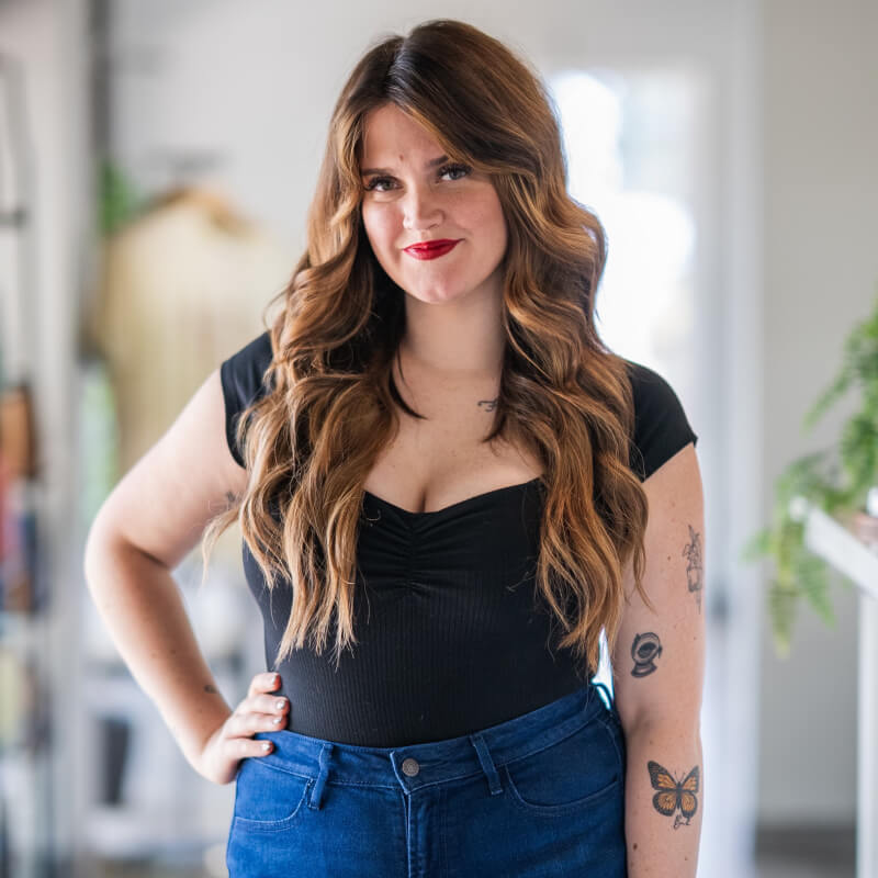 Person with long red hair sitting in a salon chair, smiling, wearing a green dress and a black belt, with salon equipment in the background. -Pure Salon, Ankeny, Johnston, & Indianola, IA
